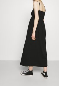 Nainen, jolla on yltäkylläinen musta midi-mekko ilman hihoja ja mustat korkeavartiset Converse-tennarit, seisoo yksinkertaista valkoista taustaa vasten.