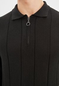 Pullover nero lavorato a maglia con chiusura a zip, dotato di colletto e texture a coste verticali. Il suo design minimalista enfatizza la semplicità.