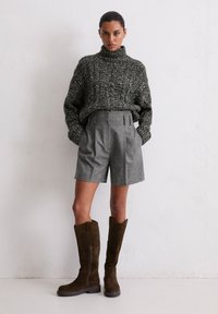 Femme portant un pull à col roulé épais gris foncé, un short gris taille haute et des bottes hautes marron foncé, se tenant contre un mur blanc.