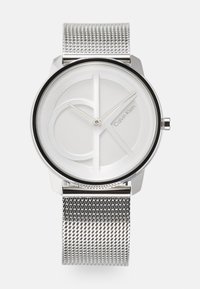Ura z rjavo mrežico blagovne znamke Calvin Klein, ki se ponaša z minimalistično belo številčnico z diskretno blagovno znamko in elegantnim črnim okvirjem. Okrogla oblika.