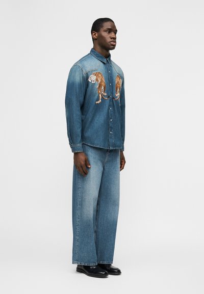 Άνδρας με μπλε τζιν πουκάμισο με κέντημα τίγρης, συνδυασμένο με φαρδιά ξεθωριασμένα μπλε τζιν και μαύρα παπούτσια, στέκεται μπροστά σε λευκό φόντο.