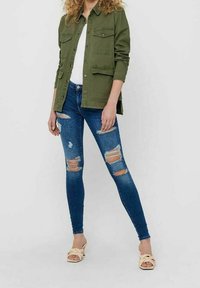 Giacca button-up verde oliva con due tasche sul petto, abbinata a jeans skinny blu strappati e sandali con tacco a blocco color crema.