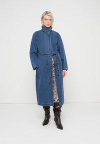 Long manteau en denim avec col montant, ceinture nouée, boutons sur le devant et poches latérales. Présente une coupe décontractée et une couleur bleue douce.