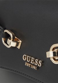 Crna torbica teksture s zlatnim akcentima. Ima logo s natpisom "GUESS EST 1981" i detalj zatvaranja nalik lancu.