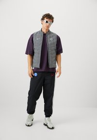 Gray quilted vest with Nike logo, purple t-shirt, black pants with blue and red patches, and light gray sneakers with textured pattern.