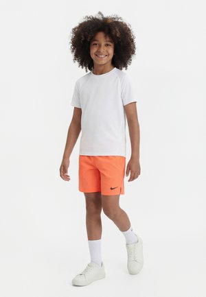 Barn med krøllet hår iført en hvid t-shirt, lyse orange Nike-shorts, hvide sokker og hvide sneakers, står og smiler.