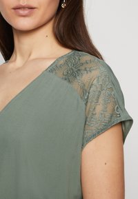 Grön blus med spetsdetaljer på axlarna. Har en v-ringning och en slät tygtextur som framhäver en avslappnad passform och elegans.