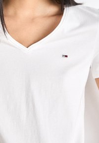 Vit bomullströja med V-ringad design. Litet broderat emblem i röd, marinblå och vit placerat på vänster bröst.