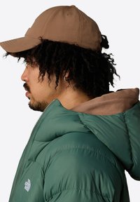 Chaqueta acolchada verde con un acabado texturizado y detalles cosidos, llevada bajo una gorra marrón con visera curvada.