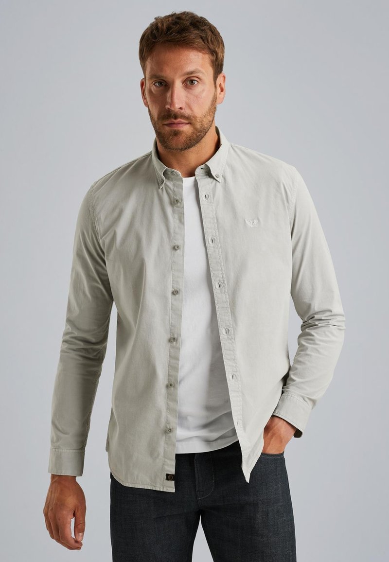 Camisa de botones de color gris claro con mangas largas, tapeta frontal y un logotipo sutil en el pecho. Llevada sobre una camiseta blanca.