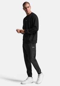 Sort sweatshirt med rund hals og logo på brystet, parret med sorte atletiske bukser med lynlåse ved anklerne, båret med hvide sneakers.
