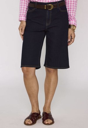 Femme portant un short en jean foncé longueur genou, ceinture marron, chemise rose à carreaux, bracelets dorés et sandales marron.