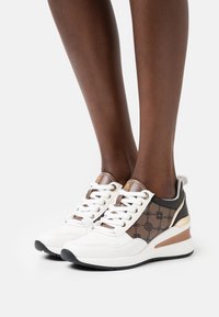 Vita och bruna sneakers med ett mönstrat sidopanel, snören, metalliska detaljer och en kraftig klack. Har en gummisula för bra grepp.