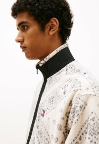 Giacca in pile di colore crema con accenti neri. Presenta un colletto alto, chiusura con zip e intricati motivi grigi sul tessuto.