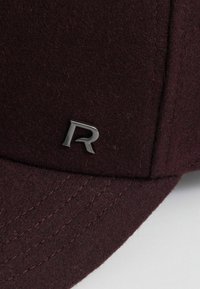 Bordowa wełniana czapka z lekko zakrzywionym daszkiem, widocznymi szwami i srebrnym metalowym logo „R” z przodu jako elementem brandingowym.