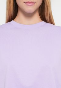 Λιλά μπλουζάκι με στρογγυλή λαιμόκοψη και κοντά μανίκια, κατασκευασμένο από απαλό βαμβακερό ύφασμα με ομαλή υφή. Χωρίς ορατά σχέδια ή διακοσμήσεις.