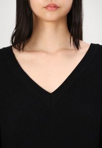 Svart strikket genser med dyp V-hals og ribbestruktur. Stoffet ser mykt ut med en subtil stretch. Passer godt til lag-på-lag styling.