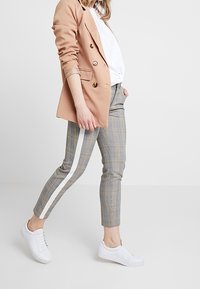 Blazer beige à fermeture à double bouton, associé à un pantalon gris à motif carreaux avec une bande latérale blanche, et des baskets blanches.
