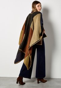 Asymmetrische poncho in een wolmix met geometrische kleurvlakken in marineblauw, bruin, groen en beige. Afgewerkt met een contrasterende stikrand.