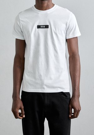 Hombre con una camiseta blanca de manga corta lisa que tiene un pequeño parche negro con el logotipo "Versace Jeans Couture" centrado en el pecho.