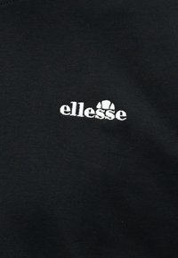 Μαύρο ύφασμα με λευκό κεντημένο λογότυπο "ellesse" που περιλαμβάνει κείμενο και ημικυκλικό σχέδιο πάνω από δύο κάθετες γραμμές.