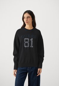 Svart sweatshirt med rund halsringning, med blå "GUESS" text och en paljettdekorering av "81" på framsidan. Långa ärmar och avslappnad passform.