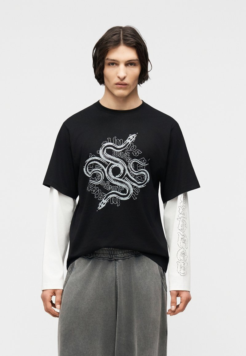 Νεαρό άτομο με μεσαίου μήκους κυματιστά μαλλιά φοράει μαύρο μπλουζάκι πάνω από λευκά μακριά μανίκια, τυπωμένο με λευκό φίδι σε σπείρα και κείμενο σε γοτθική γραμματοσειρά.