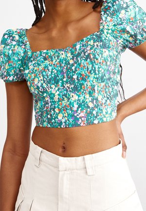 Donna che indossa un top corto verde a fiori con maniche a sbuffo e pantaloni crema a vita alta, mano appoggiata sui fianchi, torso visibile.