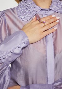 Blusa lavanda iridescente con colletto decorato da perle, polsini abbottonati e texture morbida. Mano adornata con anelli, che mostrano design intricati.