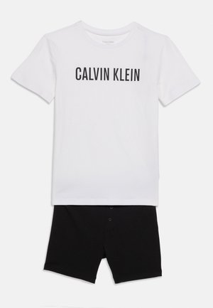 Hvid T-shirt med korte ærmer og teksten "CALVIN KLEIN" i sort over sorte shorts med knapper foran, lagt fladt på hvid baggrund.