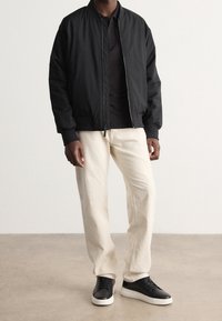 Černá bomber bunda s předním zipem, doplněná černým polokošilem, béžovými kalhotami a černými teniskami s bílou podrážkou. Hladká textura po celém povrchu.