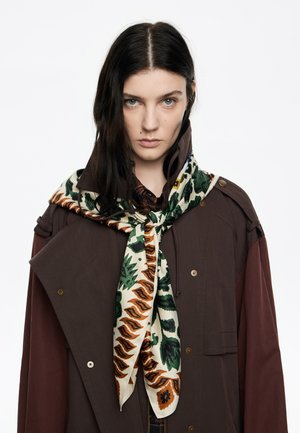 Bruine en roodachtige gelaagde buitenkleding met een decoratieve crèmekleurige en groene gestreepte sjaal die om de nek is geknoopt, met bloem- en geometrische ontwerpen.