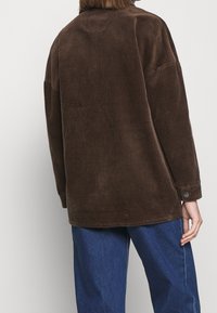 Chaqueta de pana marrón con un corte relajado, que presenta mangas largas, un cuello y detalles de botones en los puños. Tela texturizada con líneas verticales.