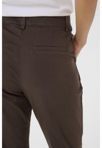 Braune Hose mit glatter Textur, ausgestattet mit zwei Gesäßtaschen, einem taillierten Bund und Gürtelschlaufen. Mittelhoher Bund.