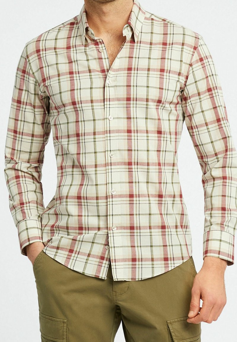 Homme portant une chemise à carreaux beige, rouge et verte à boutons, avec un pantalon cargo vert olive, debout avec une main dans la poche.