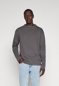 Mand iført en mørkegrå langærmet skjorte og lyseblå jeans, stående med den ene hånd i lommen mod en ensfarvet lys baggrund.