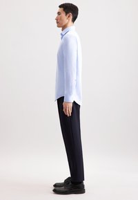 Man die in profiel staat, gekleed in een lichtblauw overhemd, zwarte broek en zwarte schoenen tegen een effen lichte achtergrond.