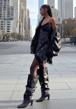 Čierne čipkované šaty s lemom s strapcami, v kombinácii s nadrozmernou čiernou koženou bundou. Celý vzhľad dopĺňajú elegantné čierne čižmy s podpätkom po kolená.
