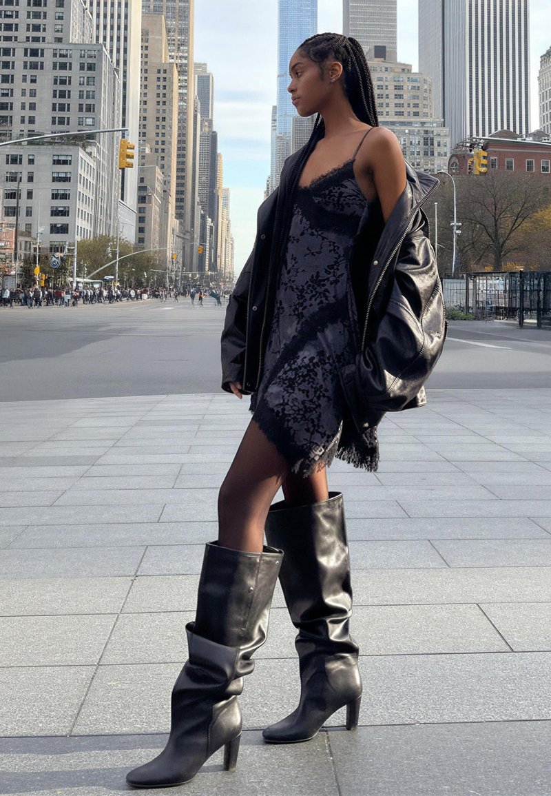 Čierne čipkované šaty s lemom s strapcami, v kombinácii s nadrozmernou čiernou koženou bundou. Celý vzhľad dopĺňajú elegantné čierne čižmy s podpätkom po kolená.