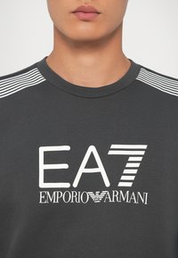 Tmavohnedá mikina s okrúhlym výstrihom, zdobená bielym textom "EA7 EMPORIO ARMANI" a pruhovanými akcentmi na pleciach. Hladká textúra.