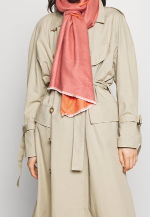 Femme portant un trench-coat beige avec une ceinture à la taille et une écharpe corail rose drapée autour du cou, debout devant un fond uni.