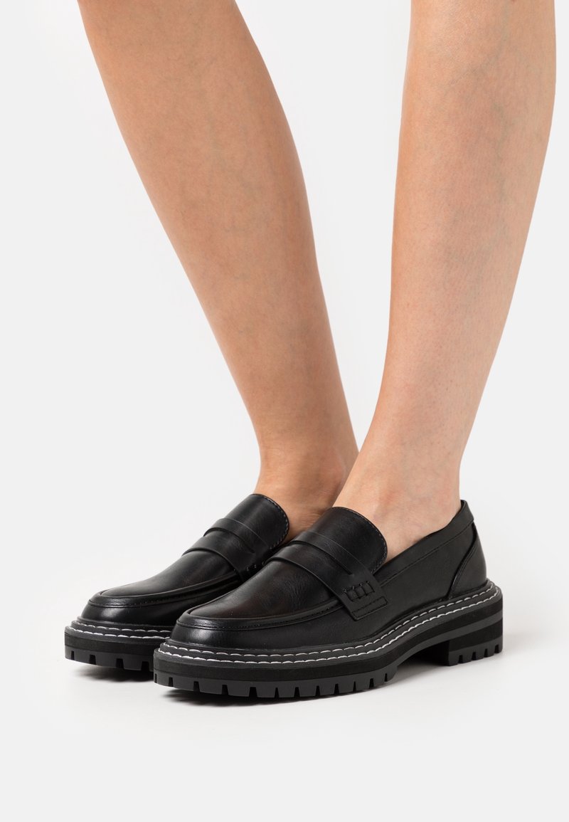 Mocassins en cuir noir avec semelles crantées épaisses et coutures blanches, portés pieds nus sur un fond uni.