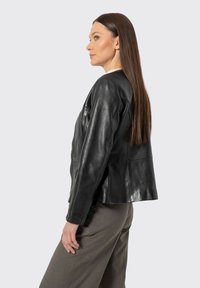 Mujer con cabello largo y liso, vestida con una chaqueta de cuero negra y pantalones grises, de pie de lado contra un fondo liso y claro.