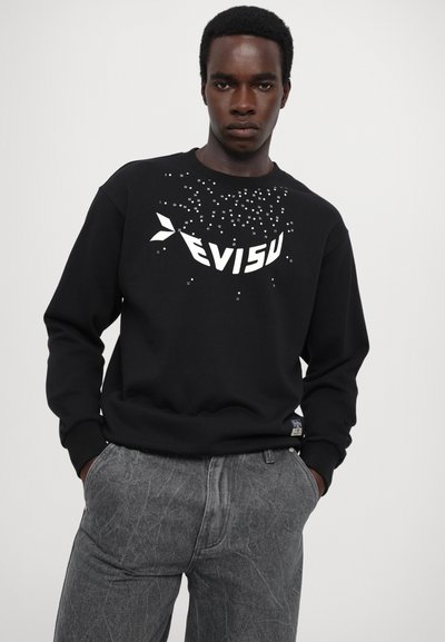 Sweatshirt noir avec un grand logo blanc "EVISU" et des motifs numériques dispersés. Manches longues et poignets côtelés. Porté avec un jean gris texturé.