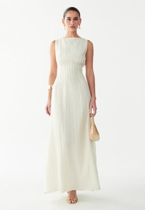 Vrouw draagt een witte mouwloze maxi-jurk, houdt een kleine beige handtas vast, gecombineerd met gouden oorbellen, armband en sandalen.