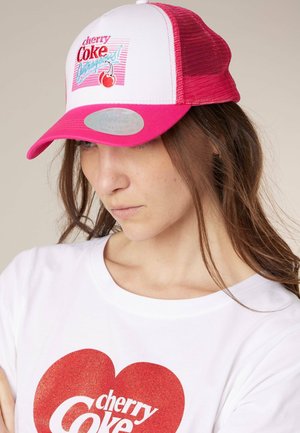 Femme portant une casquette trucker Cherry Coke blanche et rose et un t-shirt blanc avec un cœur rouge et le logo Cherry Coke.