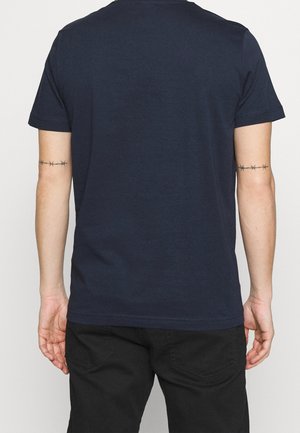 T-shirt en coton bleu marine à manches courtes et col rond, avec une texture lisse et un design simple. Un pantalon noir est partiellement visible.