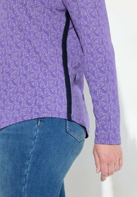Fioletowa bluzka z długim rękawem w rybi wzór, z granatową boczną wstawką i zaokrąglonym dołem. Zestawiona z niebieskimi dżinsami o dopasowanym kroju.