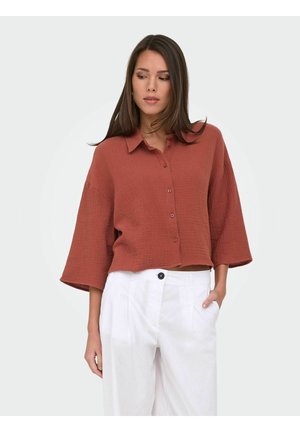 Femme portant une chemise ample couleur rouille à boutons avec de larges manches et un pantalon blanc taille haute, posant avec une main dans la poche.