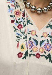Broderad krämfärgad blus med blommönster i rosa, gult, blått och grönt. V-ringad design med en mjuk, texturerad yta.